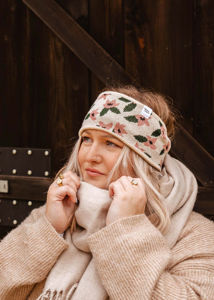 A person with light hair wears a warm and cozy Floral Blossom Headband by Mimi & August and a beige scarf, standing against a dark wooden background.