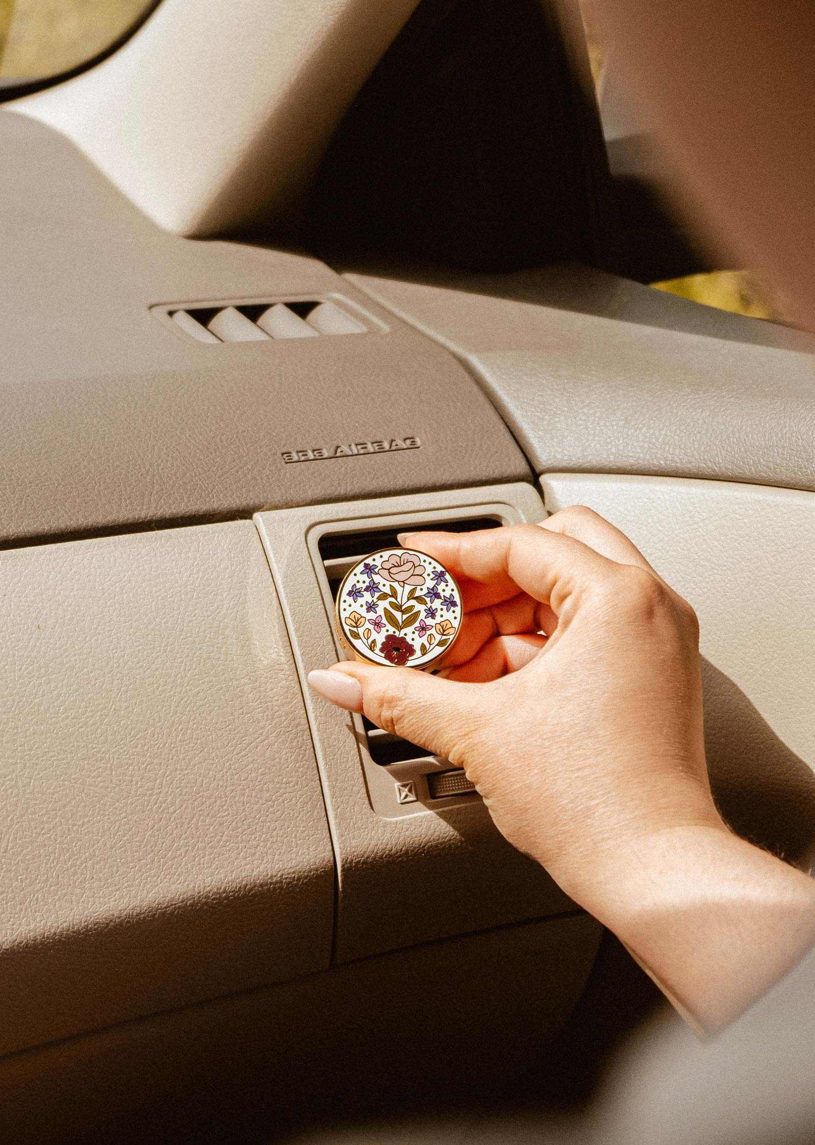Une main place le Diffuseur pour voiture fleuri de Mimi & August, au design rond floral, dans la grille d'aération d'une voiture au-dessus d'un tableau de bord beige.