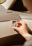 A hand places the Mimi & August Floral Car Diffuser, featuring a round floral design, into a car's air vent above a beige dashboard.