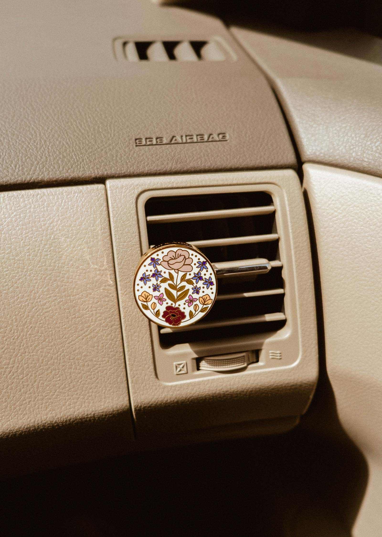 A Mimi & August Floral Car Diffuser is clipped to a car’s air vent on a beige dashboard labeled “SRS AIRBAG.”.
