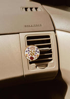 A Mimi & August Floral Car Diffuser is clipped to a car’s air vent on a beige dashboard labeled “SRS AIRBAG.”.