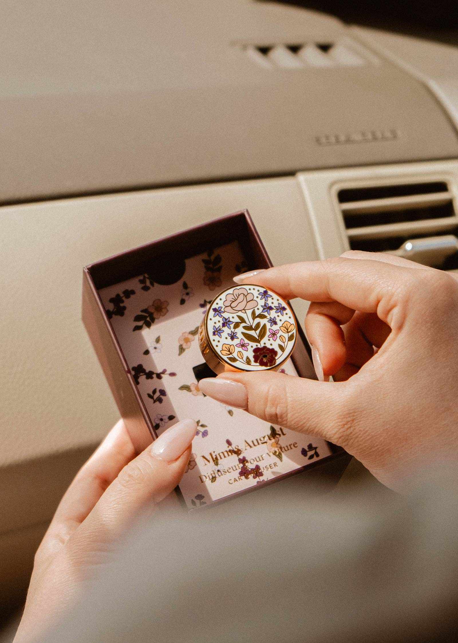 A person holds a Mimi & August Floral Car Diffuser tin above an open box with a matching floral design while sitting in a car.