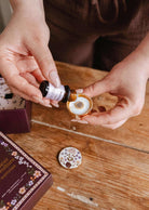 Someone pours liquid from a small brown bottle into the Mimi & August Floral Car Diffuser, a decorative round air freshener, on a wooden table beside its box and lid.