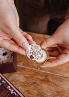 A person holds an open small round tin with a floral lid, revealing white balm inside—the Mimi & August Floral Car Diffuser that adds elegance and fragrance to your car.