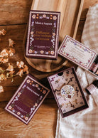 A wooden tray displays Mimi & August Floral Car Diffusers and scented candles, accented with small flowers, on a wooden table draped with a checkered cloth.