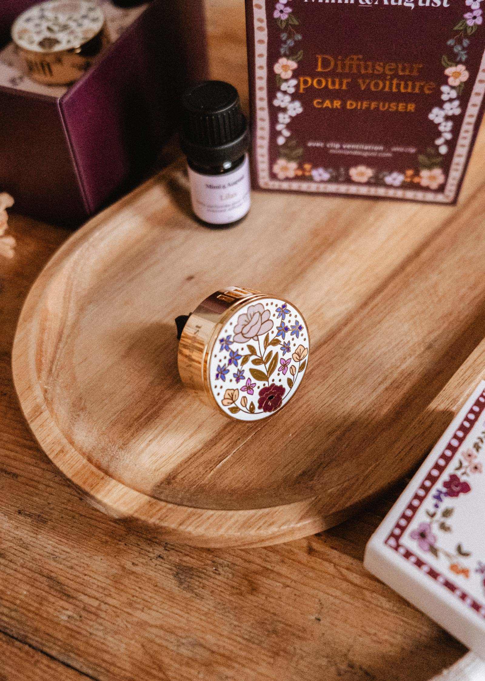 Le Diffuseur pour voiture fleuri de Mimi & August, au design rond floral, est présenté sur une assiette en bois à côté d'une bouteille d'huile parfumée et d'une boîte étiquetée « Diffuseur pour voiture ».
