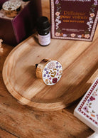 The Mimi & August Floral Car Diffuser, featuring a round floral design, is displayed on a wooden plate next to an essential oil bottle and a box labeled "Diffuser pour voiture.