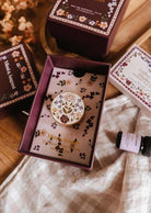 The Mimi & August Floral Car Diffuser is a decorative floral enamel pin, showcased in an open purple box among similar boxes and a glass vial on wood—ideal as an enamel car diffuser or unique car air freshener accent.