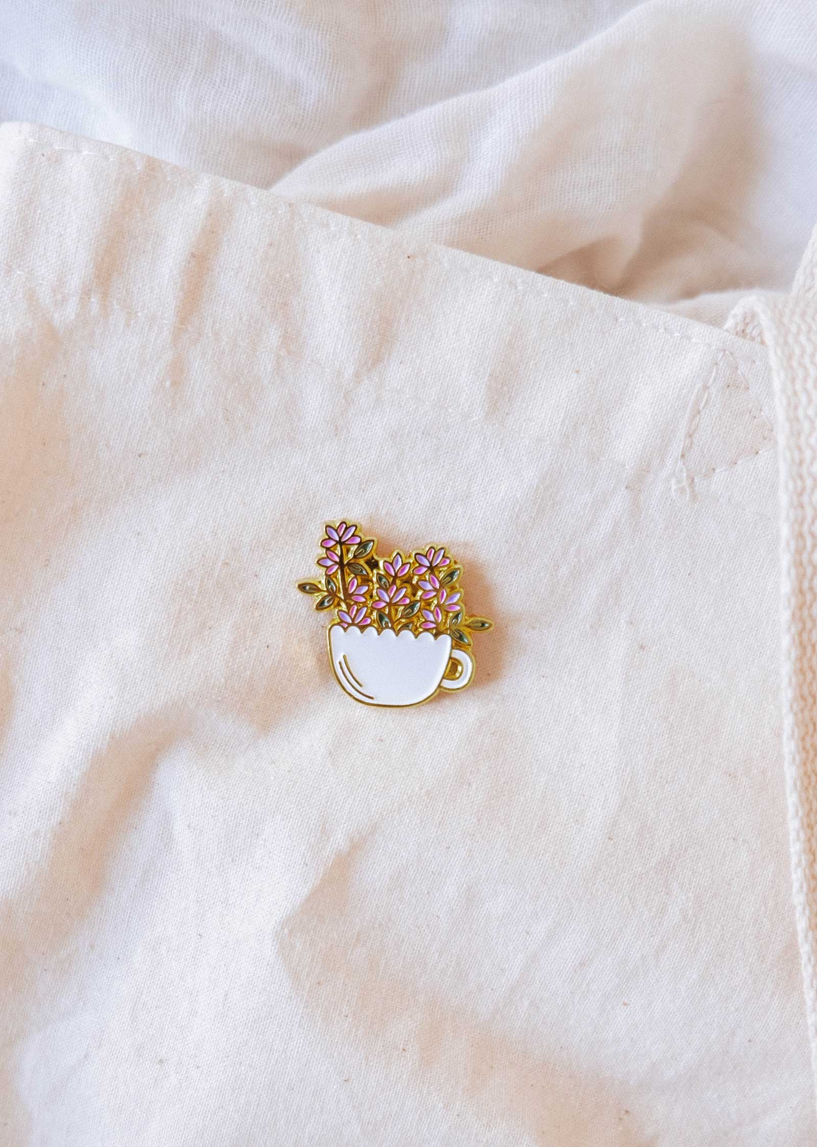 The Mimi & August Bloom Cup Enamel Pin, shaped like a white teacup with purple and yellow flowers, is attached to a cream-colored fabric bag.