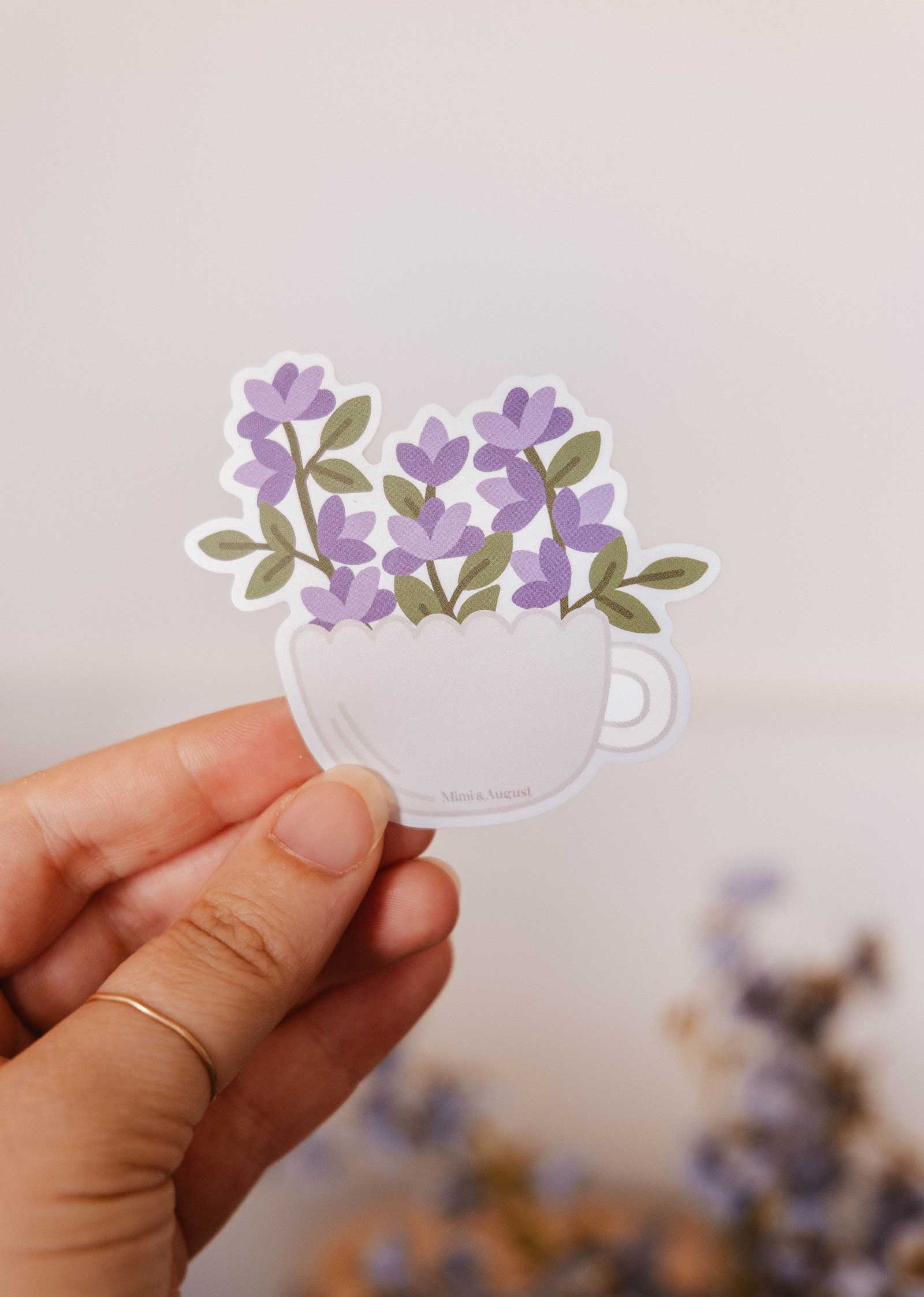A hand holds the Bloom Cup - Vinyl Sticker by Mimi & August, showing a white teacup filled with purple flowers—perfect for cute stationery lovers—set against a neutral background.