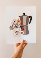 Une main tenant une illustration d'une cafetière et des fleurs mimi et august.