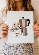 Une femme brandit une affiche avec une cafetière italienne et des fleurs de la marque mimi et august.