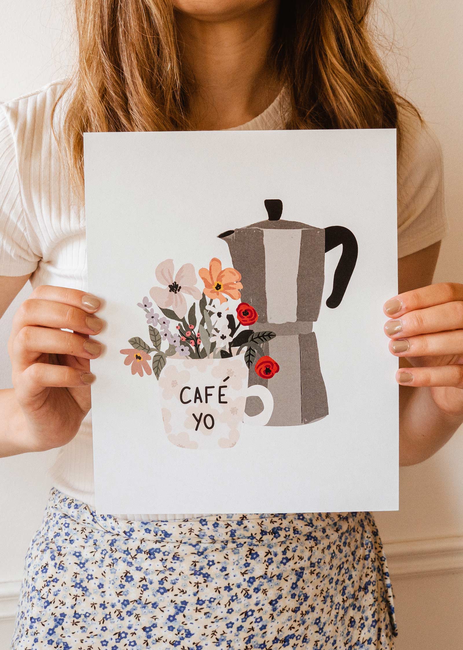 Une femme brandit une affiche avec une cafetière italienne et des fleurs de la marque mimi et august.