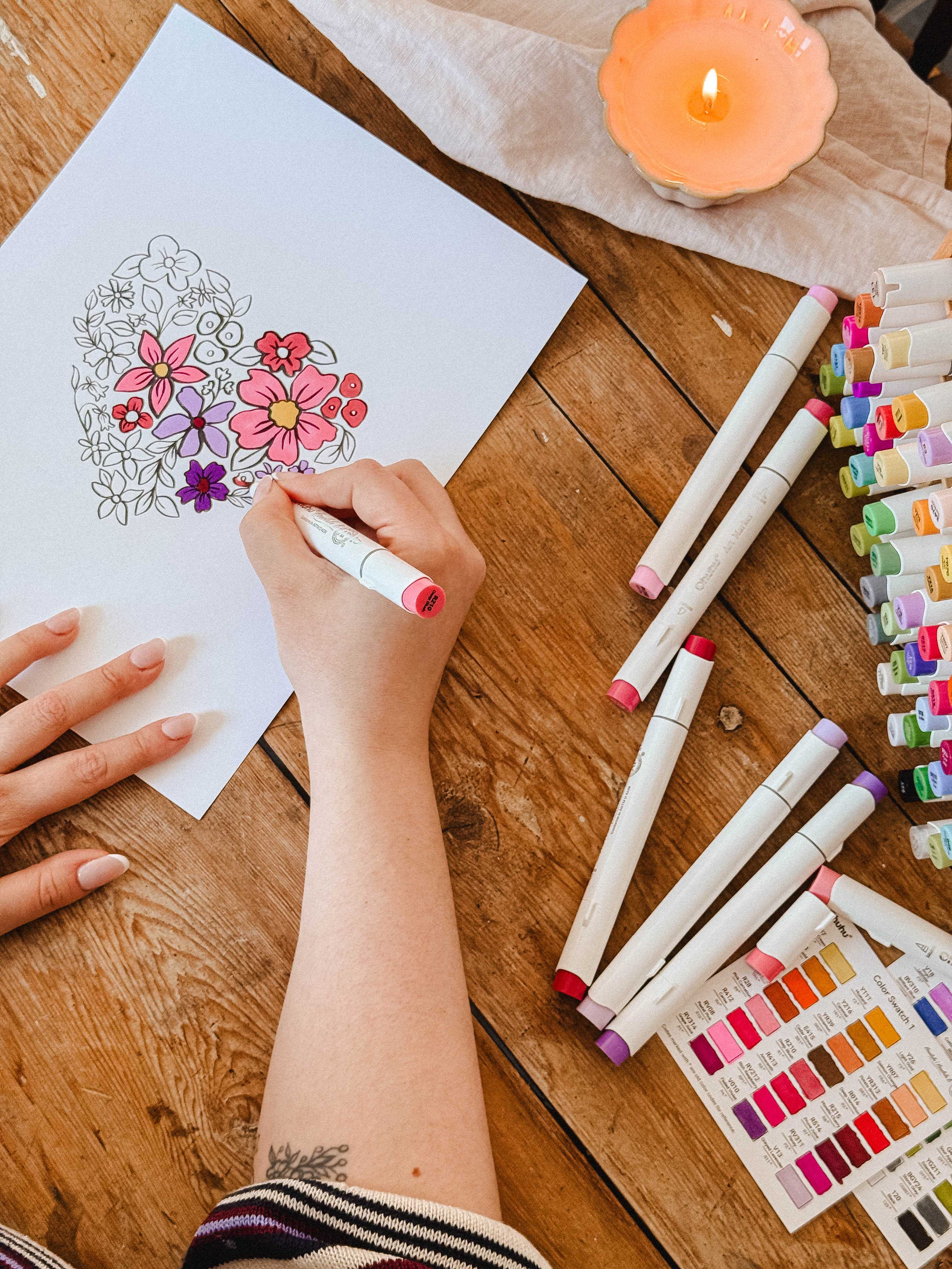 A person colors the Mimi & August Floral Heart Coloring Sheet with markers, surrounded by colorful markers, a color chart, and a lit candle—a perfect printable or free digital download for your next relaxing art session.