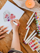 A person colors the Mimi & August Floral Heart Coloring Sheet with markers, surrounded by colorful markers, a color chart, and a lit candle—a perfect printable or free digital download for your next relaxing art session.