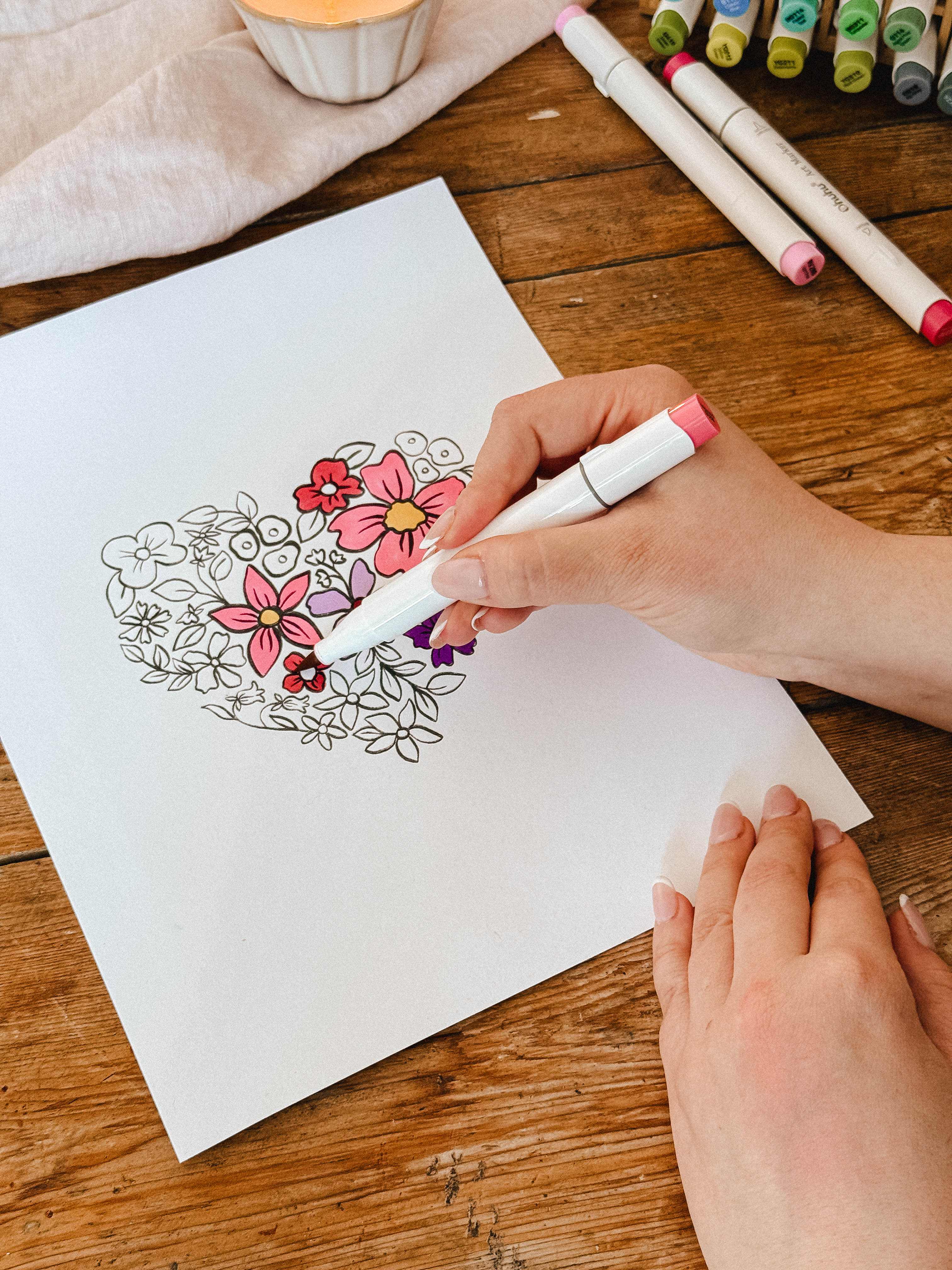 Someone colors the Mimi & August Floral Heart Coloring Sheet on white paper with pink and purple markers at a wooden table, enjoying a relaxing moment with this free coloring sheet.