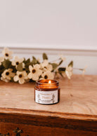 A 2 oz Flores soy candle by Mimi & August, in a small amber jar, is lit on a wooden surface with white flowers in the background. Hand-poured in Montreal.