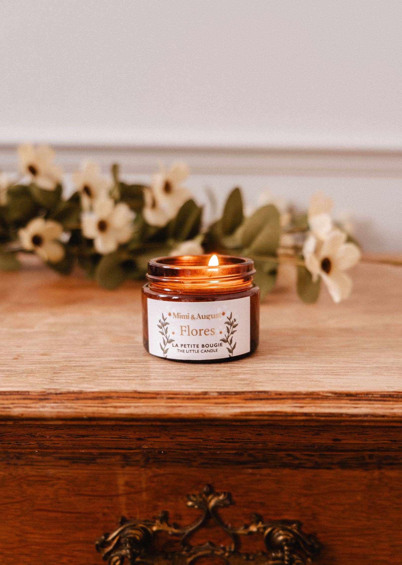 A lit Flores – 2 oz Soy Candle by Mimi & August, hand-poured in Montreal, sits in a brown glass jar on a wooden surface with blurred cream artificial flowers in the background.