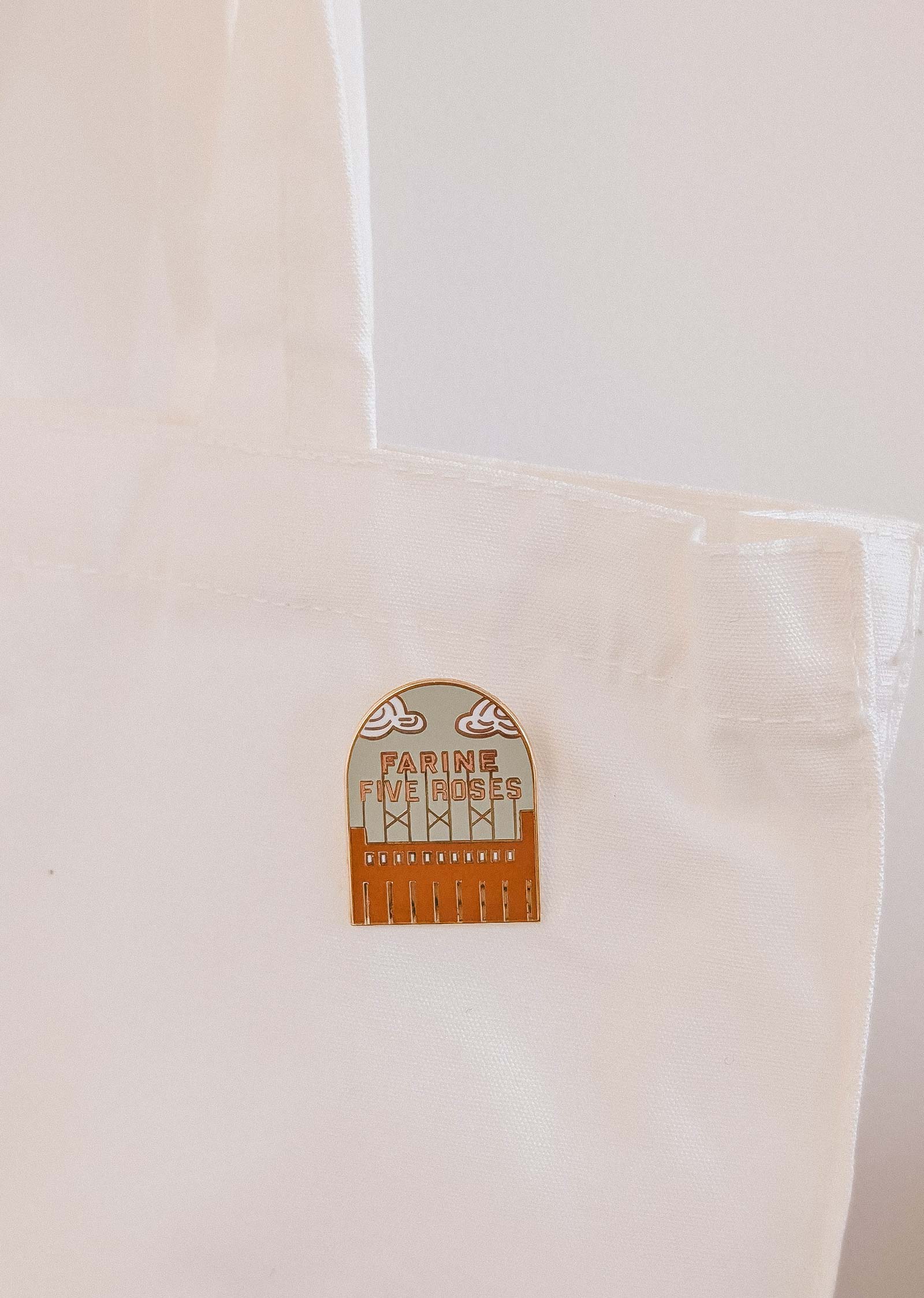 The Flour & Roses Factory Enamel Pin by Mimi & August is attached to the handle of a white fabric tote bag, adding a retro Montreal-inspired accent.