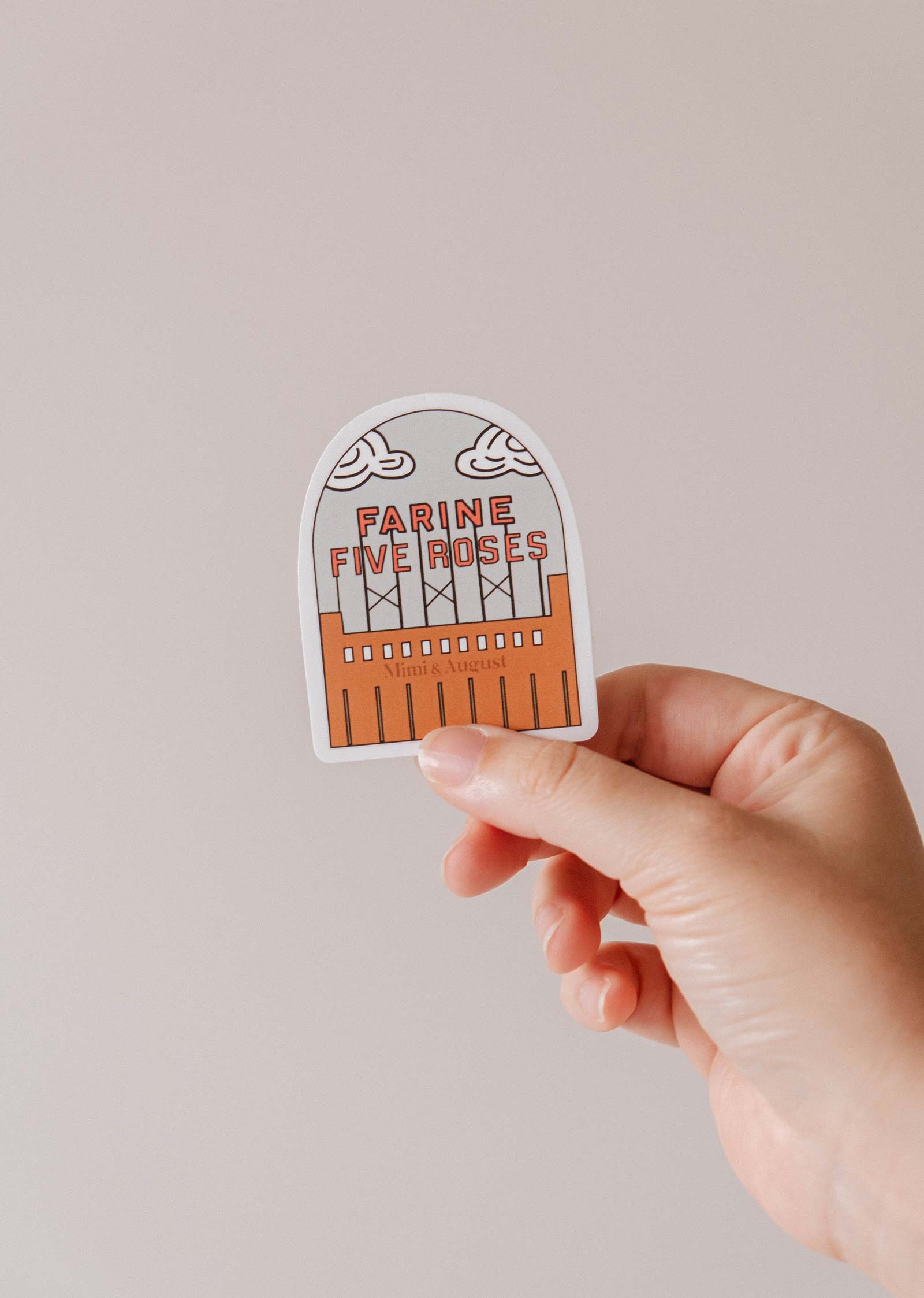 A hand holds Mimi & August’s Flour & Roses Factory vinyl sticker, featuring a retro illustration of Montréal’s Farine Five Roses sign with clouds and an orange building—an essential for architecture fans.