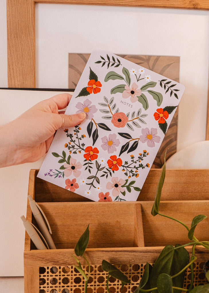 Une main tient un carnet Mimi & August Bombe de fleurs, orné d'un motif floral, illustrant la créativité florissante et l'amour des fleurs.
