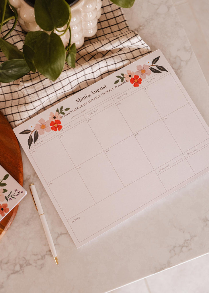 Un Planificateur Semainier Bombe de Fleurs de Mimi & August sur une table à côté d'une plante et d'un stylo.