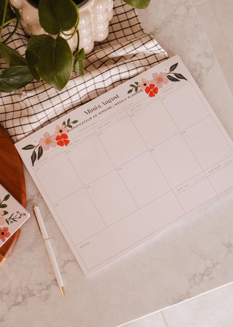 An eco-friendly Flower Bomb - Weekly Planner by Mimi & August on a table next to a plant and a pen.