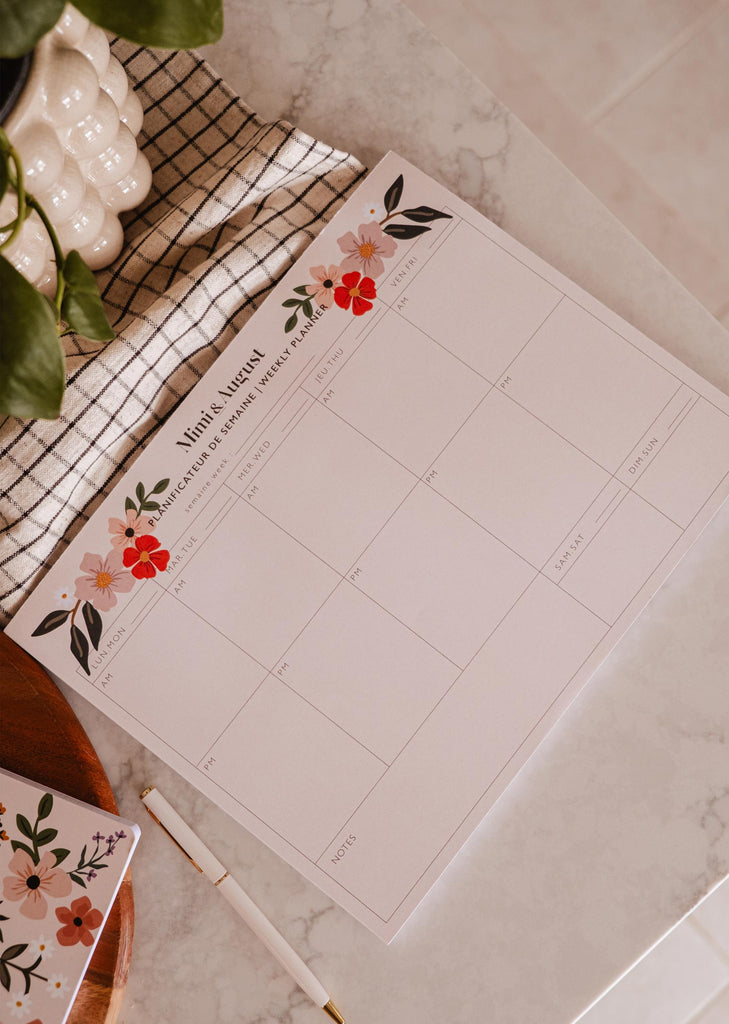 Un Planificateur Semainier Bombe de Fleurs orné de belles fleurs, posé sur une table à côté d'une plante en pot. (Marque : Mimi & August)