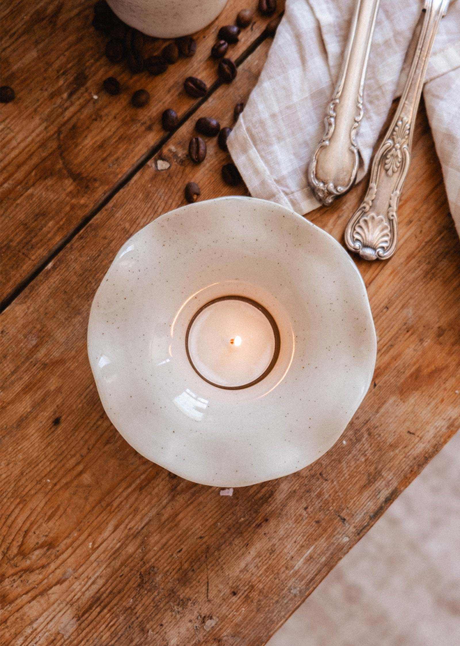 A lit tealight candle in the Fluted Ceramic Tealight Holder by Mimi & August rests on a wooden table beside antique cutlery, a white cloth, and scattered coffee beans.