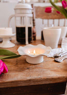Une bougie allumée dans le Bougeoir en céramique cannelé de Mimi & August repose sur une table en bois avec une cafetière à piston, des tasses, des ustensiles et des tulipes roses.