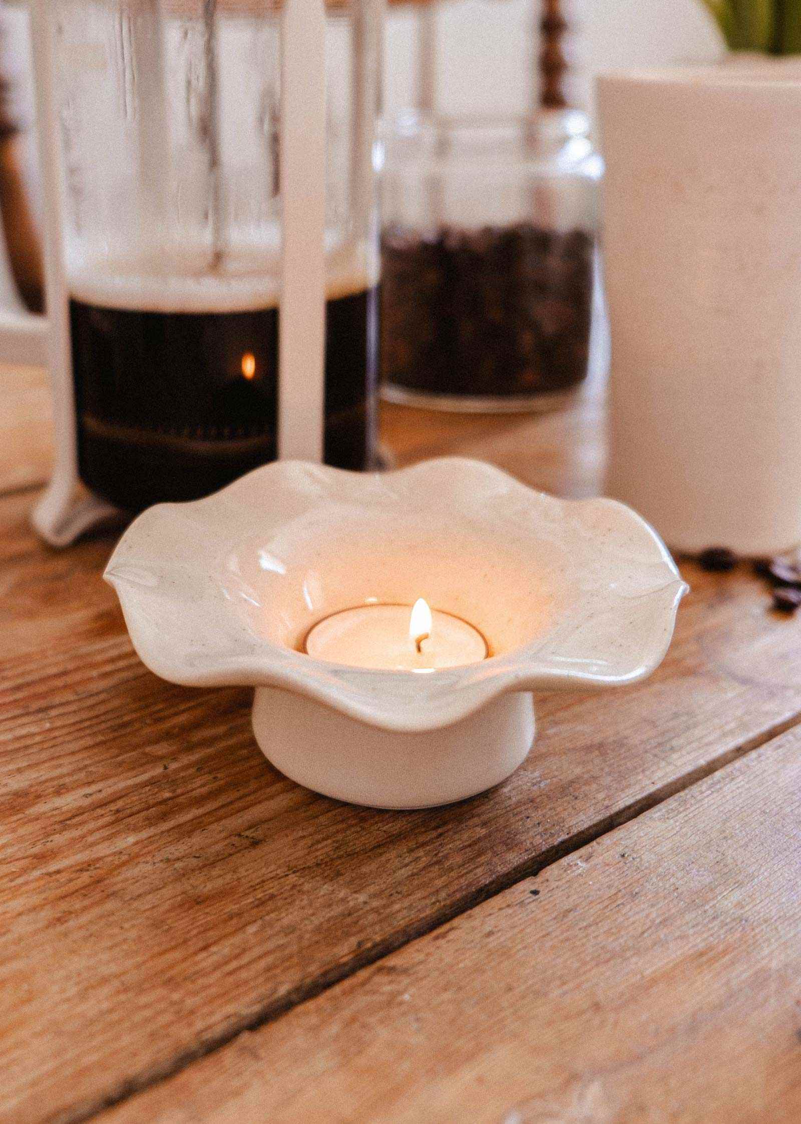 A lit tealight candle in the Fluted Ceramic Tealight Holder by Mimi & August sits on a wooden table, with a French press, a jar, and a cup in the background.