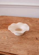 The Fluted Ceramic Tealight Holder by Mimi & August, featuring a wavy-edged white ceramic design, sits on a wooden table against a plain background.