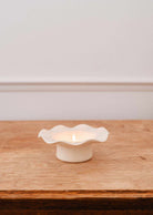 Une bougie brille dans le Bougeoir en céramique cannelé de Mimi & August, son design blanc cannelé aux rebords ondulés présenté sur une surface en bois contre un mur blanc.