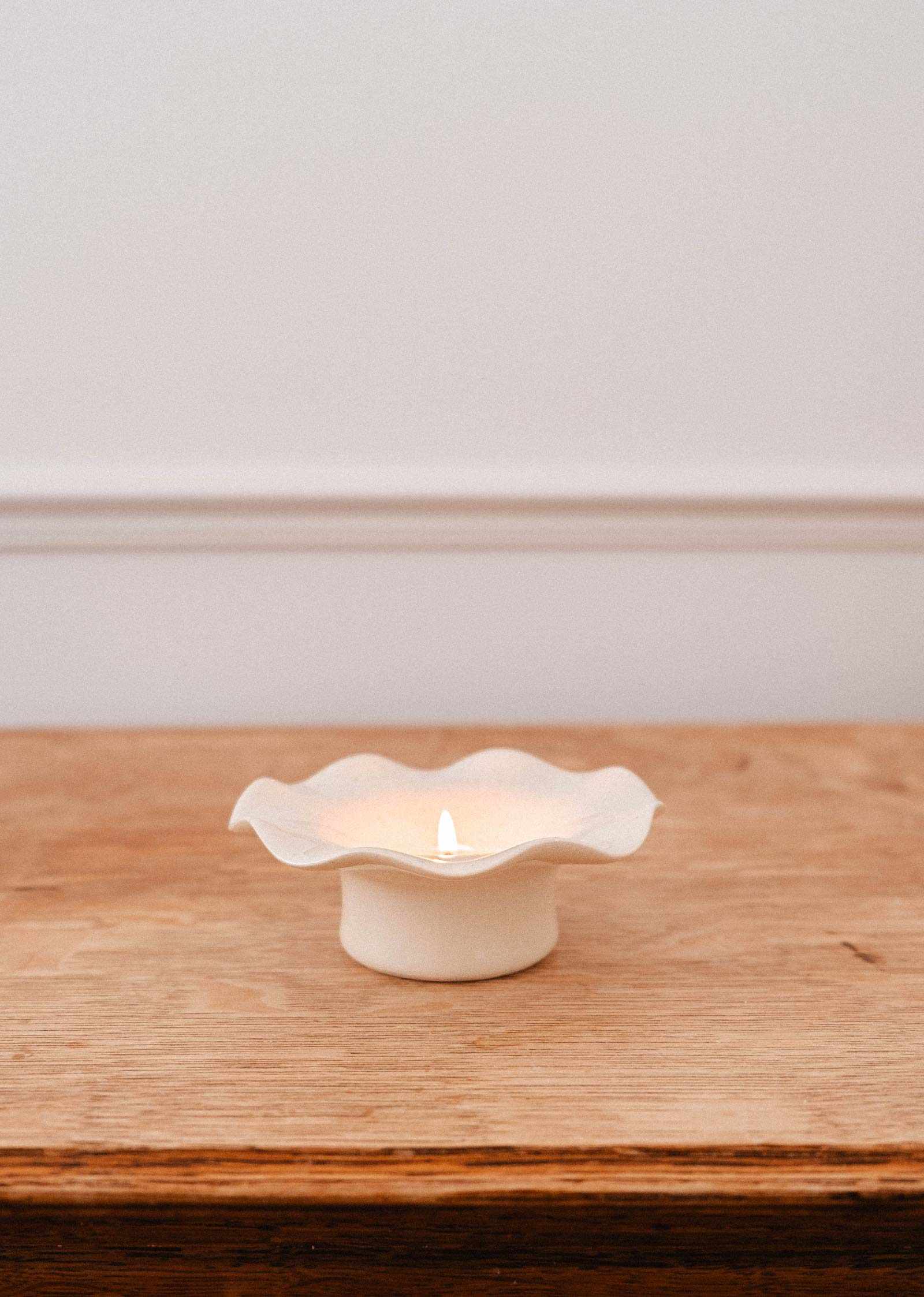 Une bougie brille dans le Bougeoir en céramique cannelé de Mimi & August, son design blanc cannelé aux rebords ondulés présenté sur une surface en bois contre un mur blanc.