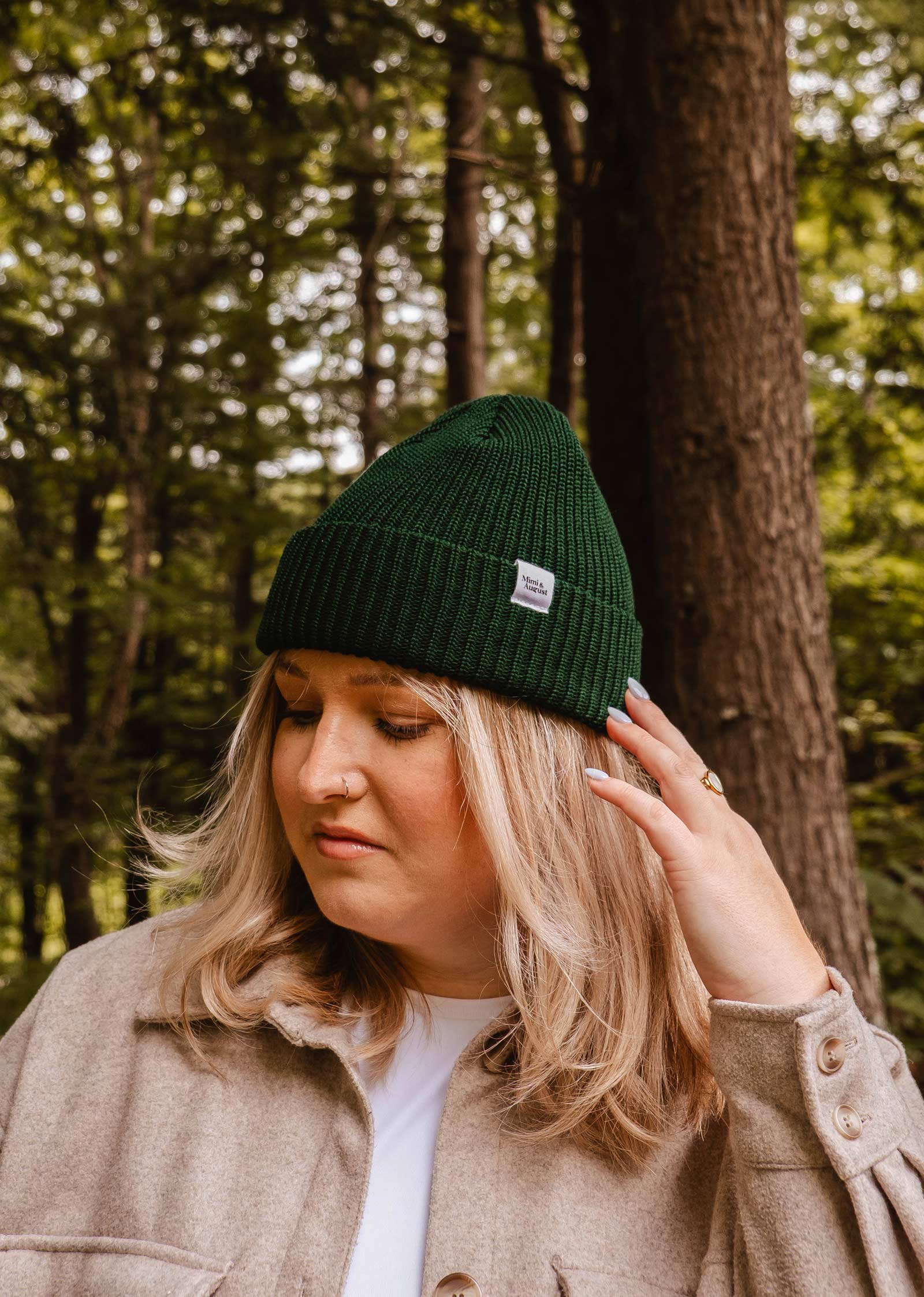 Une femme aux cheveux clairs, vêtue d'un bonnet Forest Green Cap Super Puff Beanie de Mimi & August et d'une veste beige, se tient dans une forêt luxuriante, se touchant le côté de la tête.