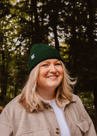 Une personne aux cheveux blonds, portant un bonnet Mimi & August Forest Green Cap Super Puff Beanie et un manteau beige, sourit alors qu'elle se tient à l'extérieur dans une forêt.