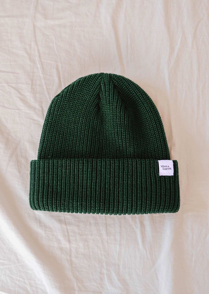 A Forest Green Cap Super Puff Beanie from Mimi & August, crafted from plush yarn and featuring a small white label, lies on a cream-colored textured fabric surface—a winter must-have.