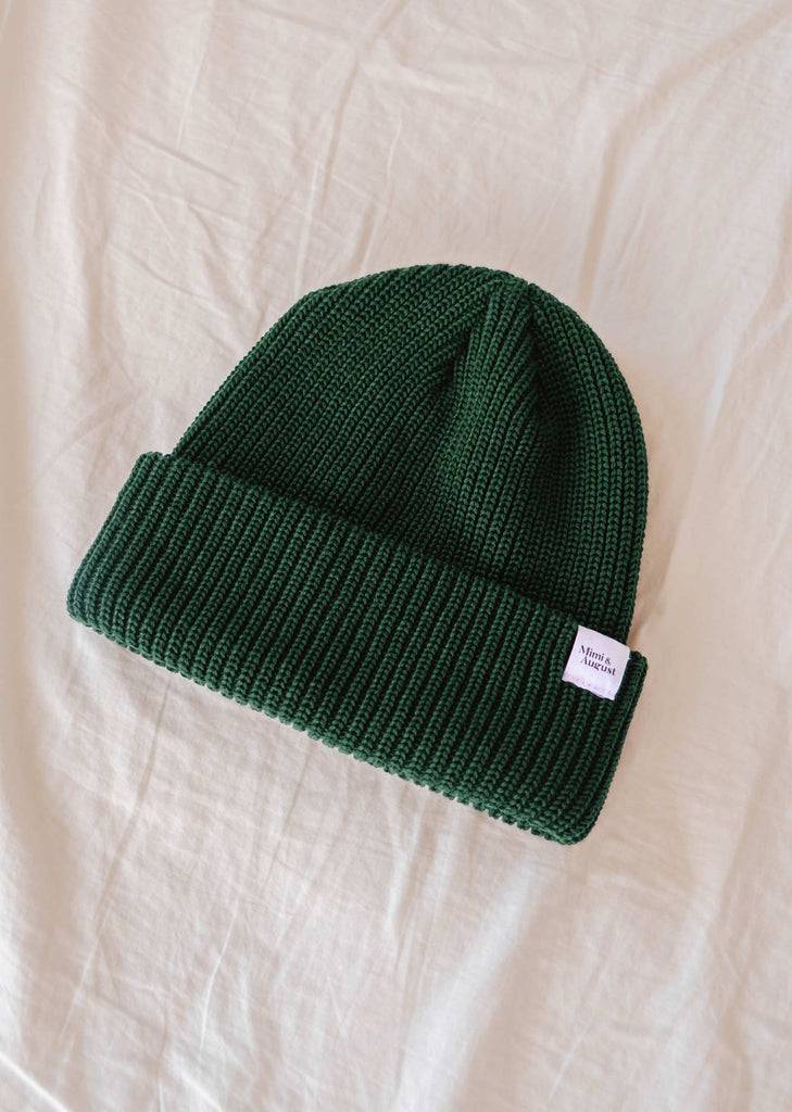 A Forest Green Cap Super Puff Beanie by Mimi & August, made from plush yarn and featuring a small white label on the fold, is laid on a wrinkled beige fabric surface, making it a winter must-have.