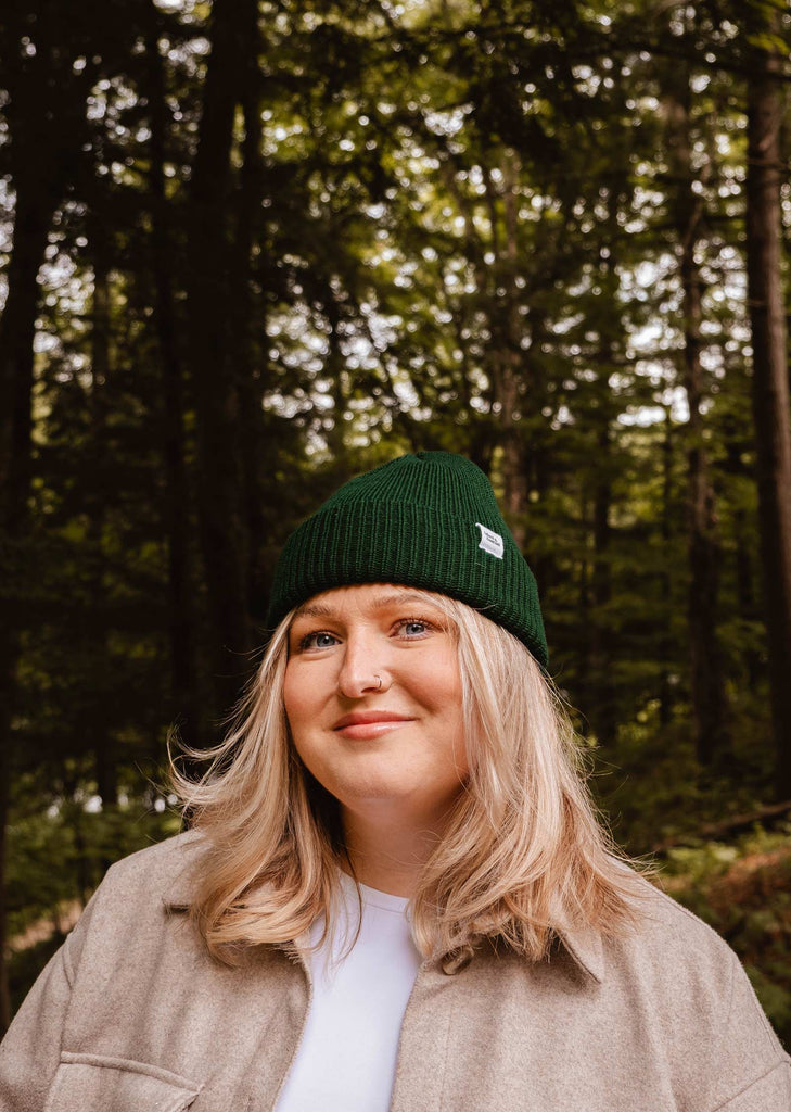 A woman with long blonde hair wears a Forest Green Cap Super Puff Beanie by Mimi & August and a beige coat, standing in a forested area with dense trees in the background. This winter must-have, crafted from plush yarn, perfectly complements her serene surroundings.