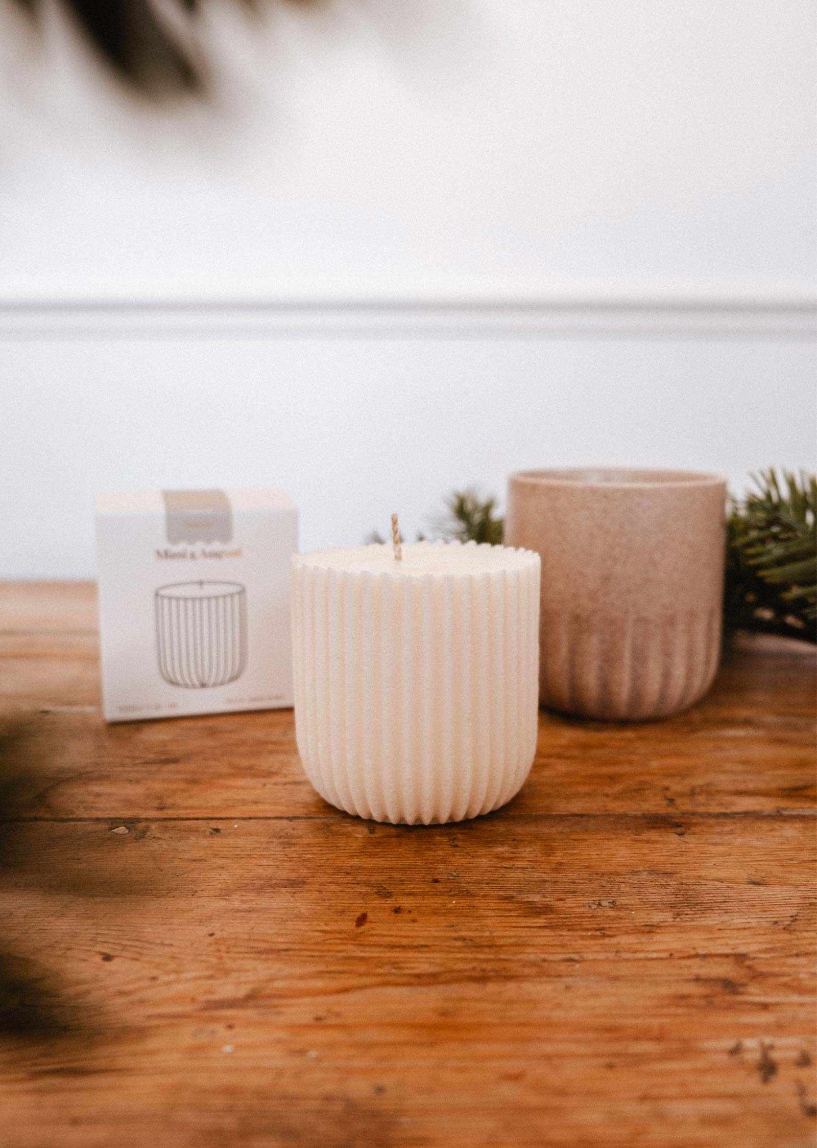The Candle Refill Foresta by Mimi & August, a ribbed white soy wax candle, rests on a wooden table near a brown cup, small box, and evergreen branches.