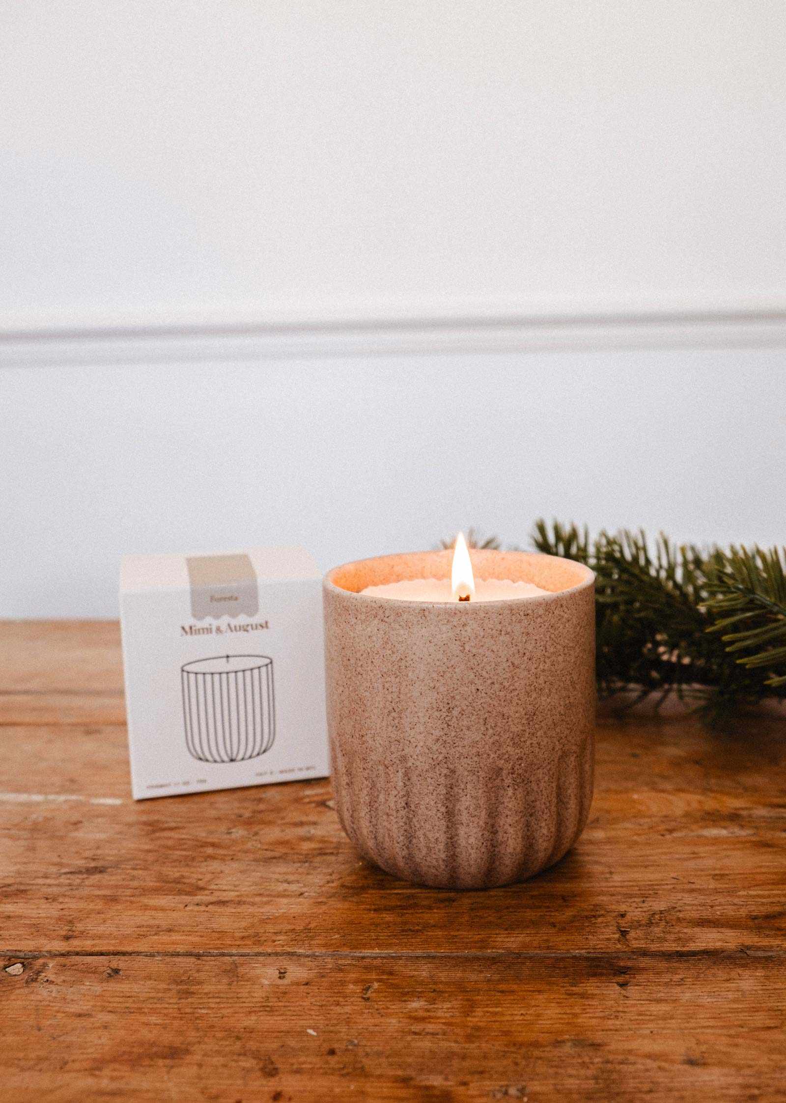 The Mimi & August Candle Refill Foresta, made with soy wax, sits in a textured beige container on a wooden surface beside its box and greenery, releasing a subtle fir tree scent into the air.