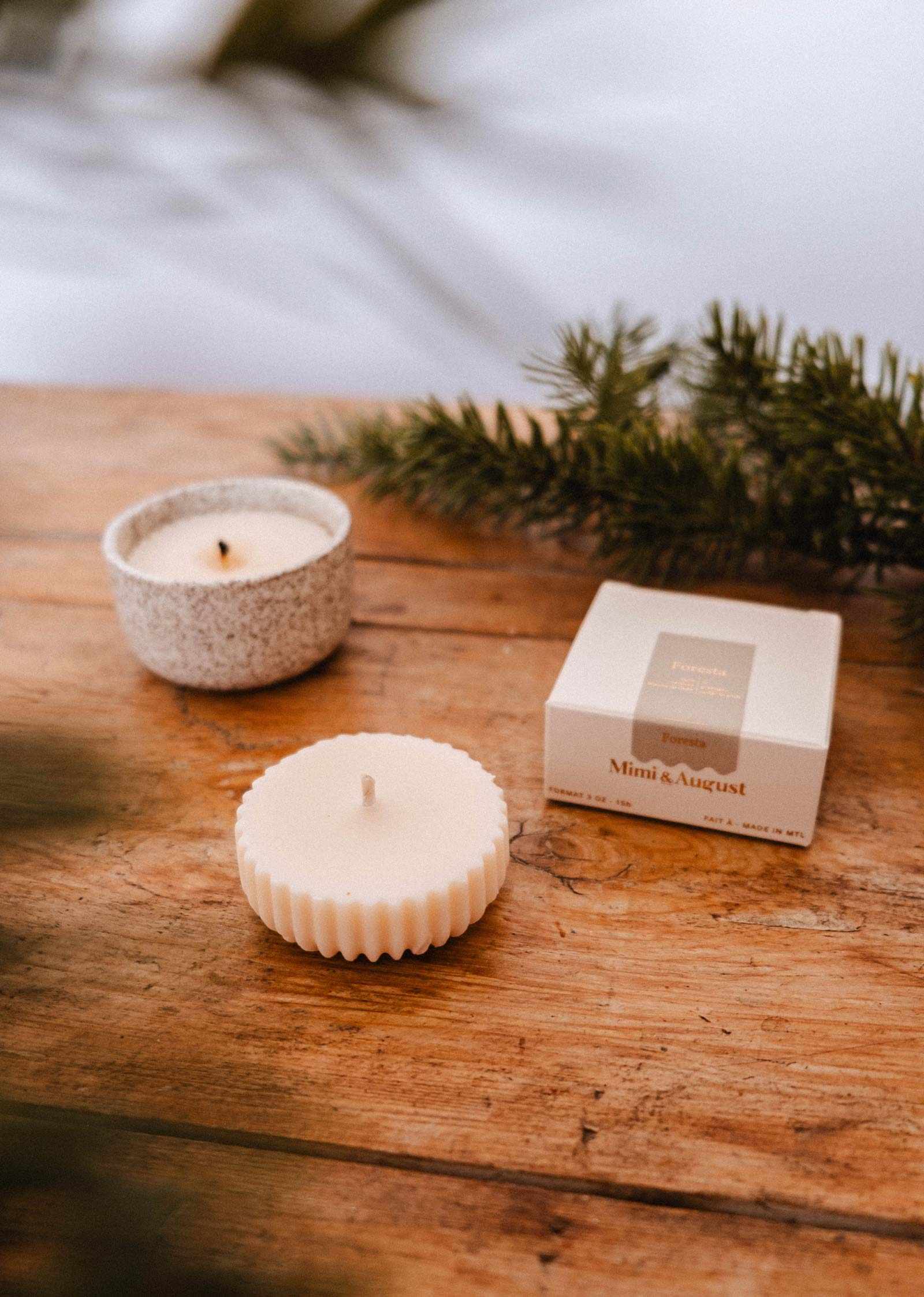 Mimi & August’s Candle Refill Foresta, a white soy wax candle in a textured dish with fluted edges and a labeled box, is displayed on wood with pine branches, capturing the fresh fir tree aroma.
