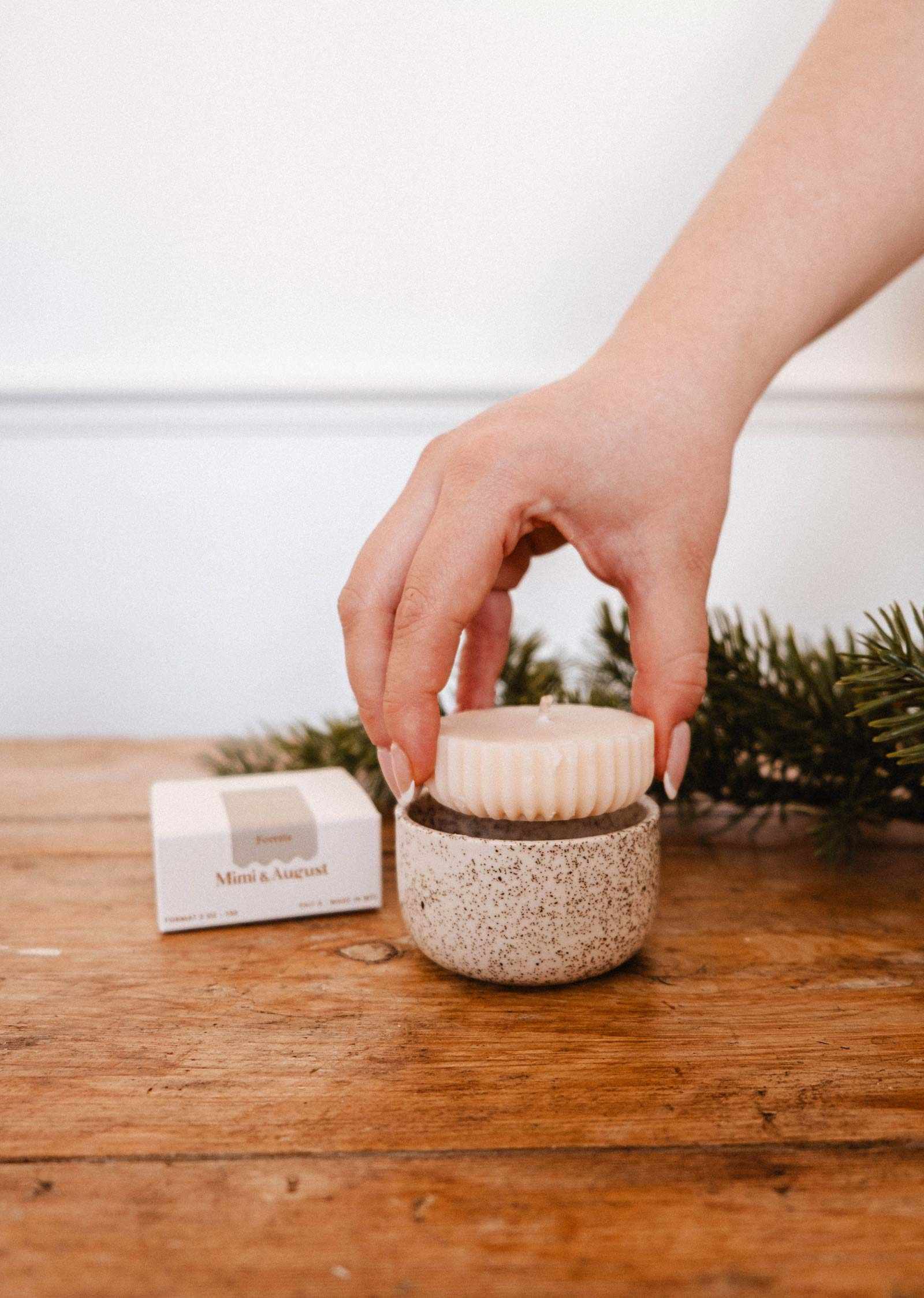 A hand places a round soap bar on a speckled ceramic container, with the Mimi & August Candle Refill Foresta and a pine branch on a wooden surface in the background.