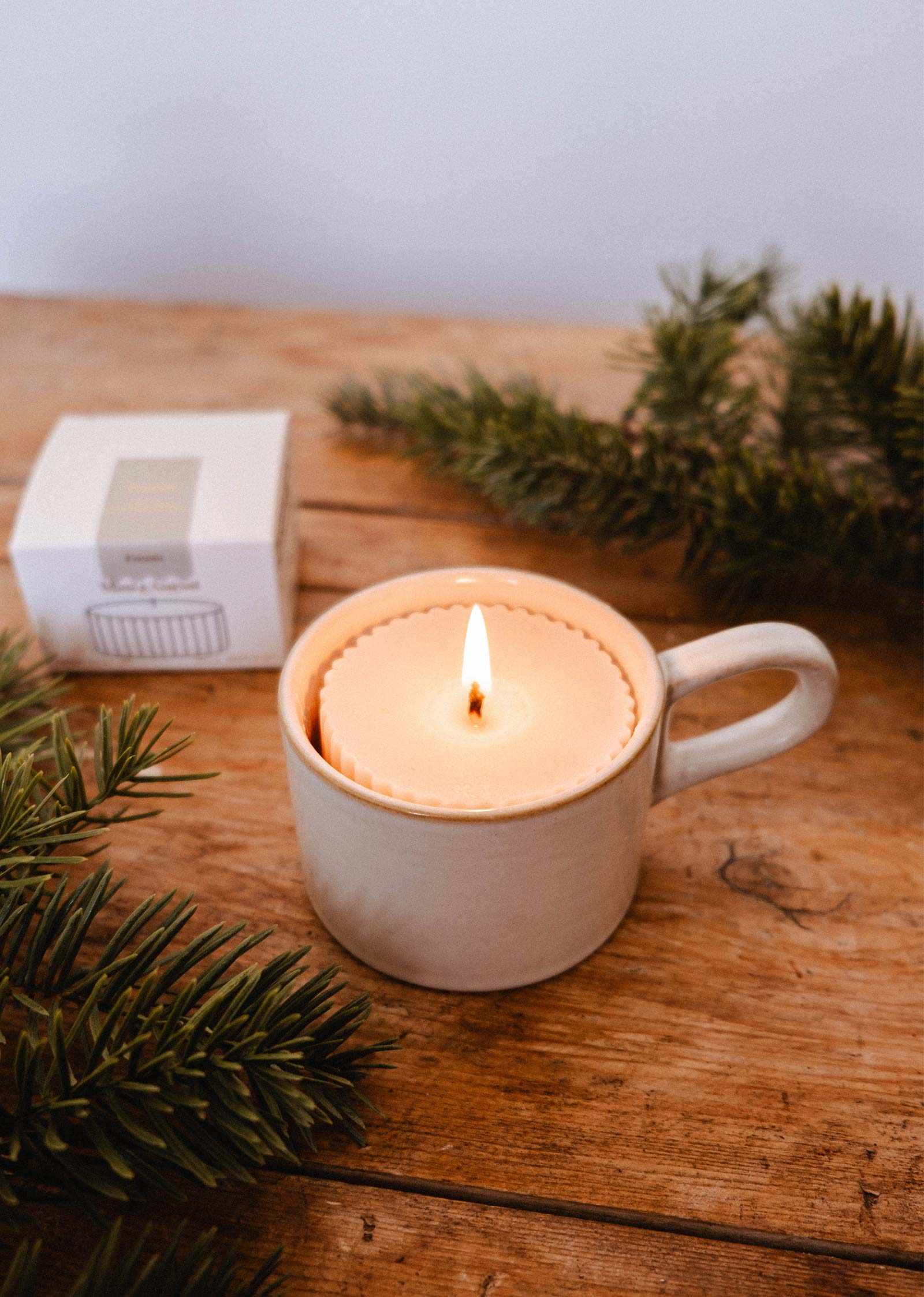 The Candle Refill Foresta by Mimi & August, a soy wax candle with a soft fir tree aroma, rests in a ceramic mug on wood among pine branches and a white box.