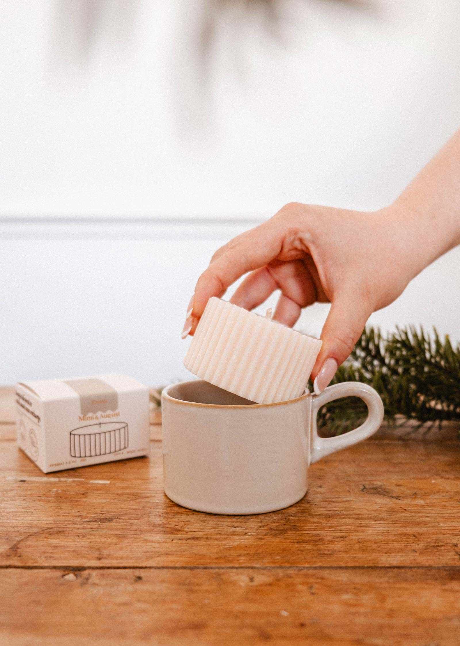 A hand places the Candle Refill Foresta by Mimi & August into a beige ceramic mug on a wooden table, with its box and a pine branch in the background.