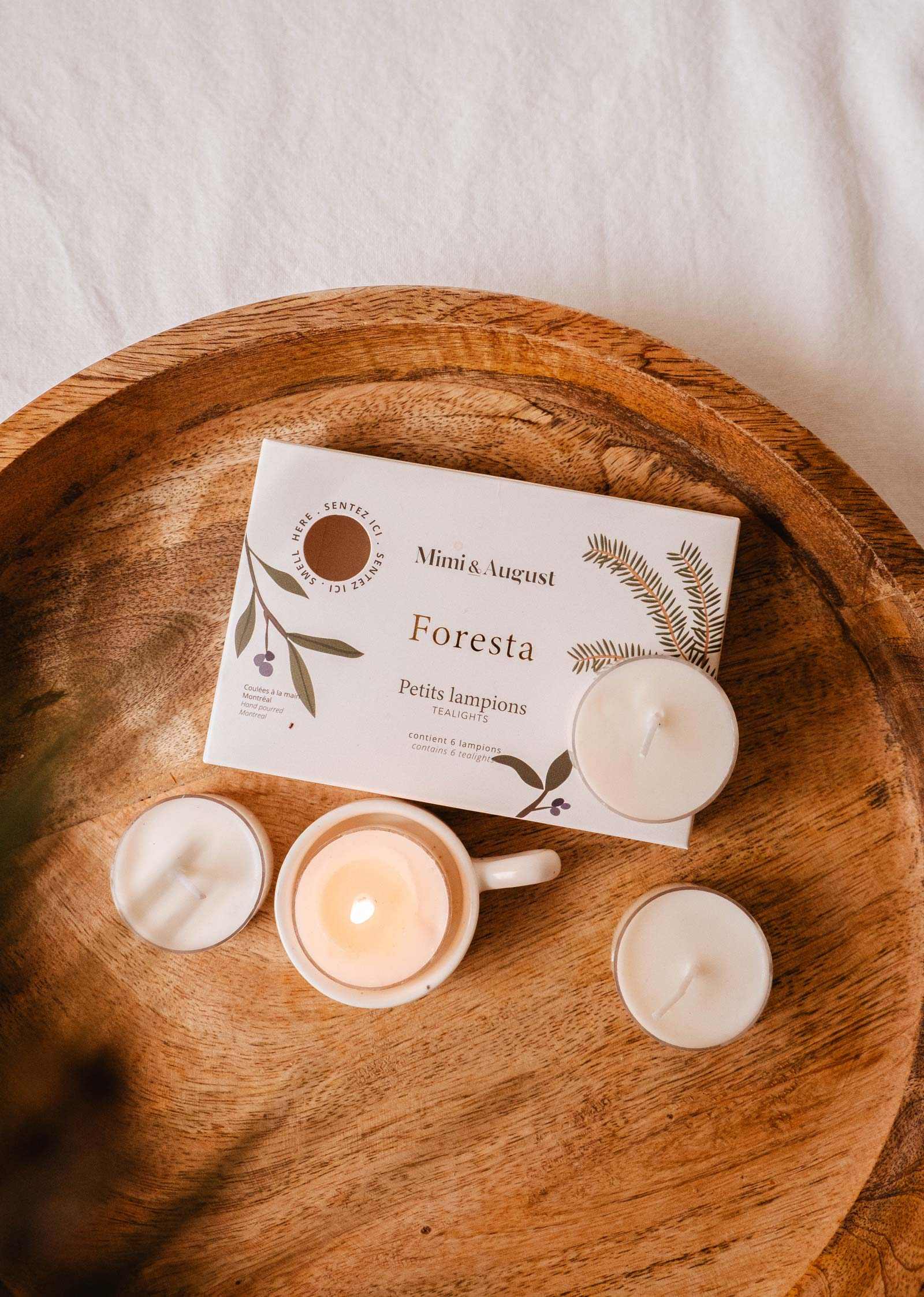 The Mimi & August Foresta Tealight Candle Set includes four white soy wax tealights in a wooden tray and a Foresta box, perfect for adding a forest-inspired fragrance to your home.