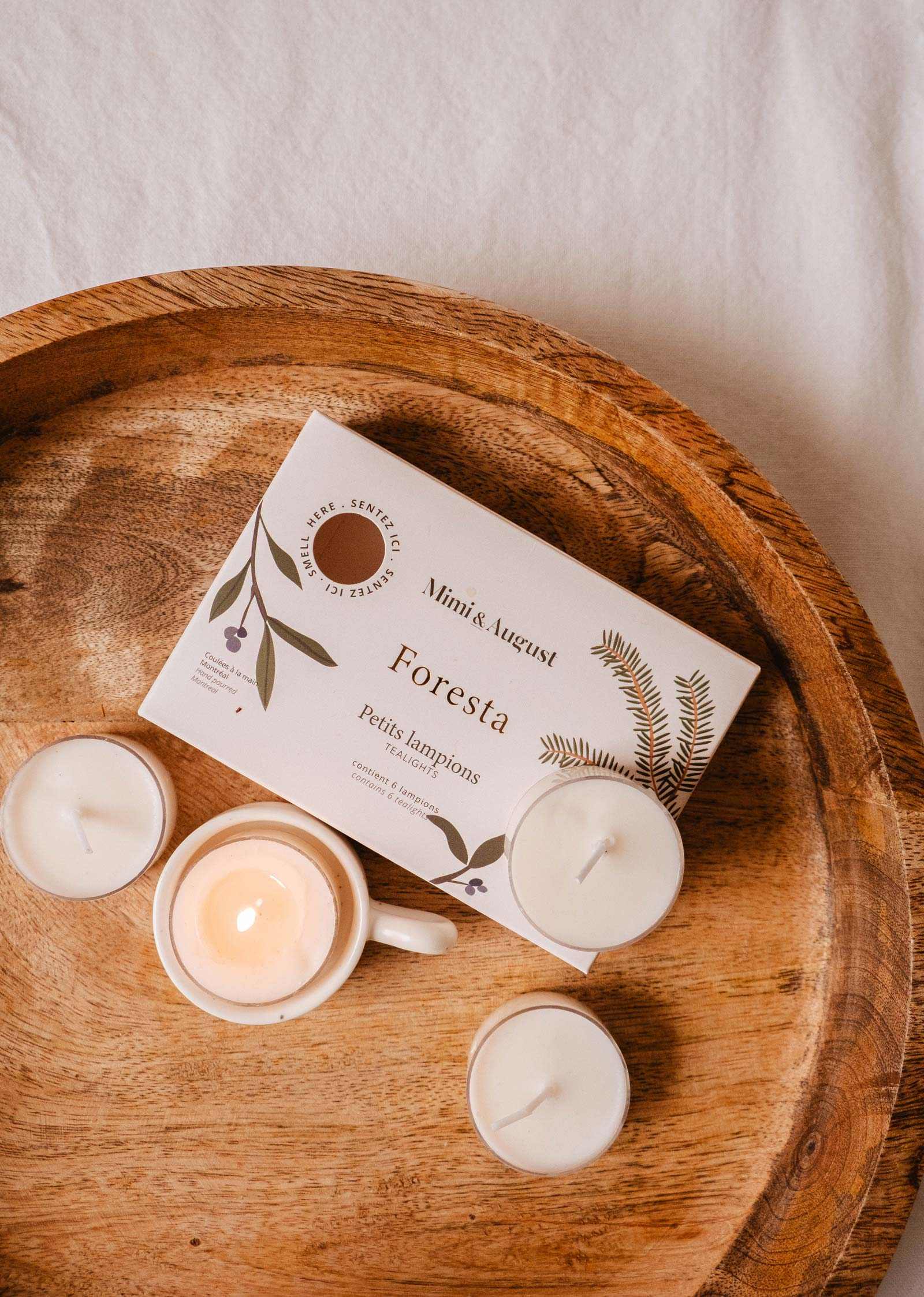 A wooden tray holds a Mimi & August Foresta Tealight Candle Set with botanical designs, featuring a white candle in a cup and three soy wax tealights.