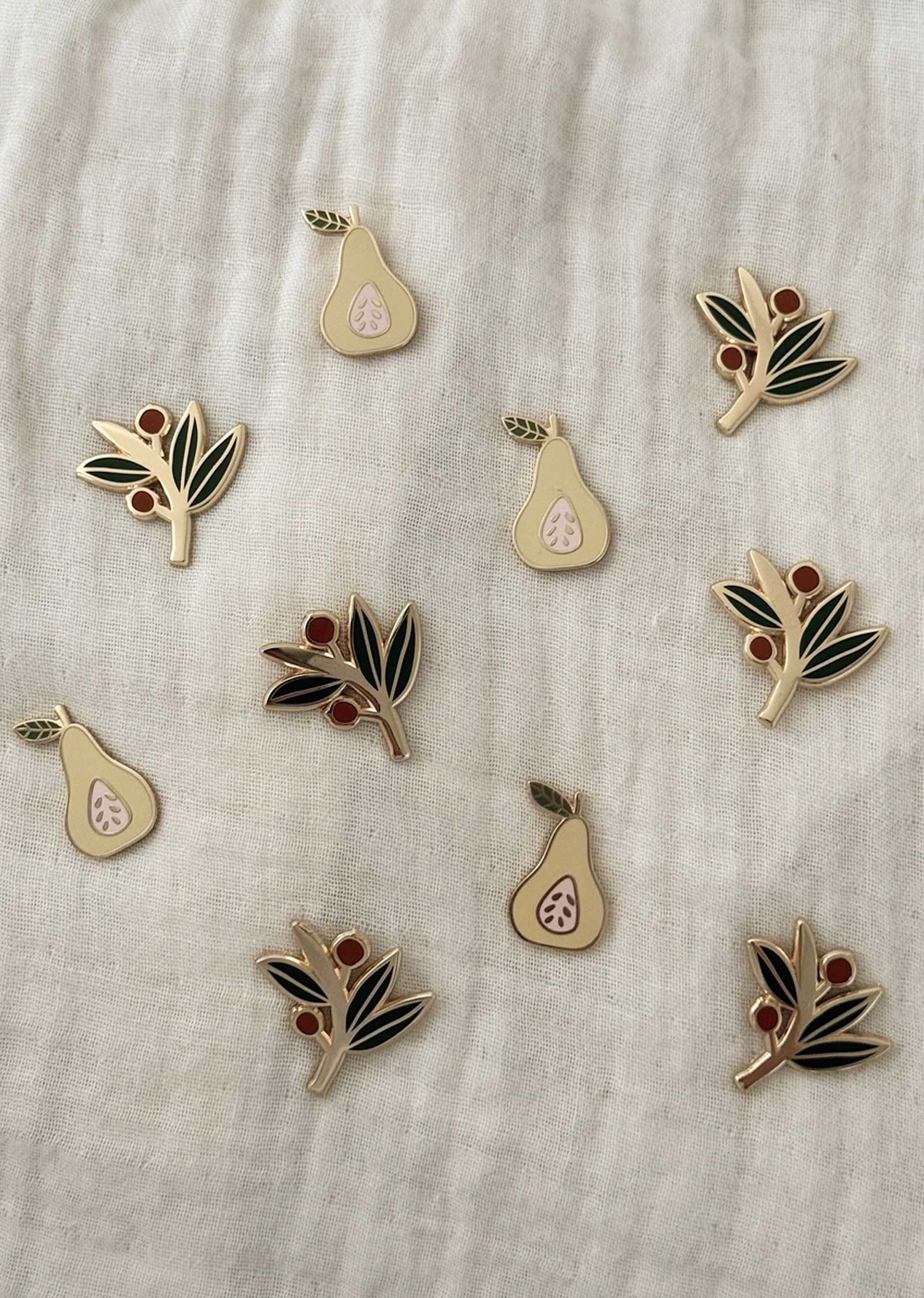Ten pear and olive branch enamel pins from Mimi & August are displayed on a light fabric background—perfect for your collection or as a gift. Get them as Free Enamel Pins (100% off) in the BOGO Bundle at BOGOS.io.