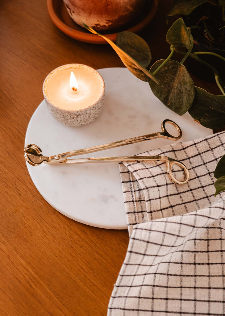 A durable Mimi & August stainless steel wick trimmer next to a reusable candle on a wooden table.