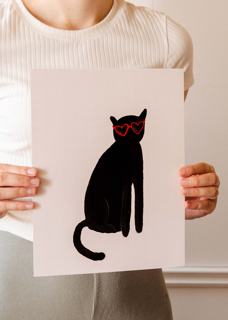 A woman holding up a Mimi & August Heart Black Cat Art Print wearing heart-shaped sunglasses.